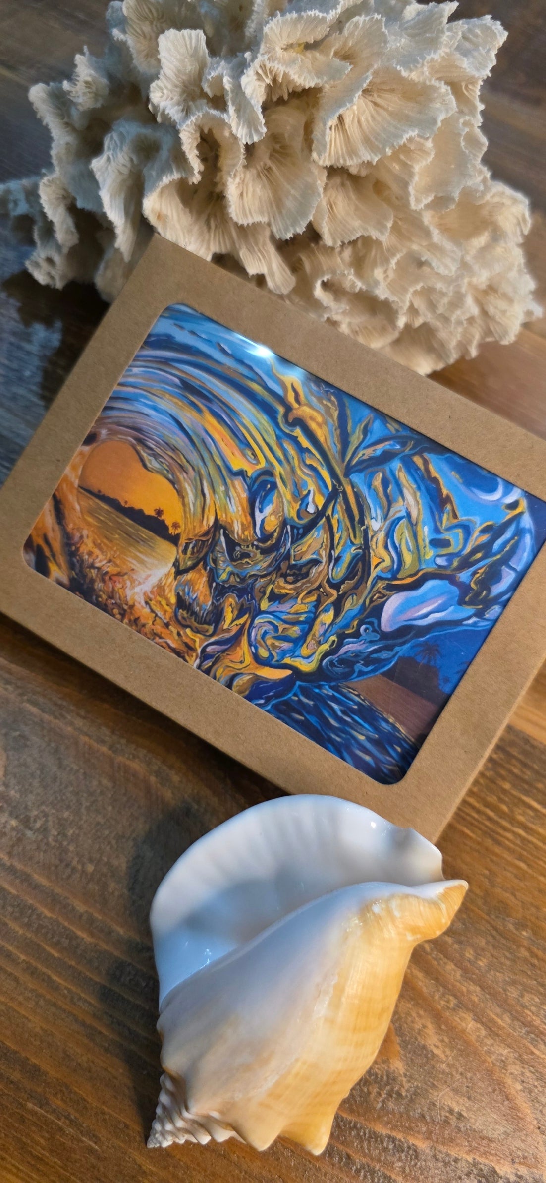 A square art print or small framed artwork with an abstract blue, yellow, and orange scene, placed on a wooden surface with a large seashell in front and coral-like decor behind it.