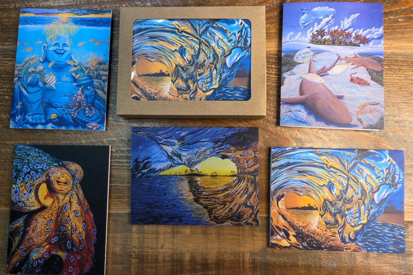 Five colorful square art prints or cards arranged on a wooden tabletop, each showing a different abstract landscape or scene in blue, gold, orange, and black tones.