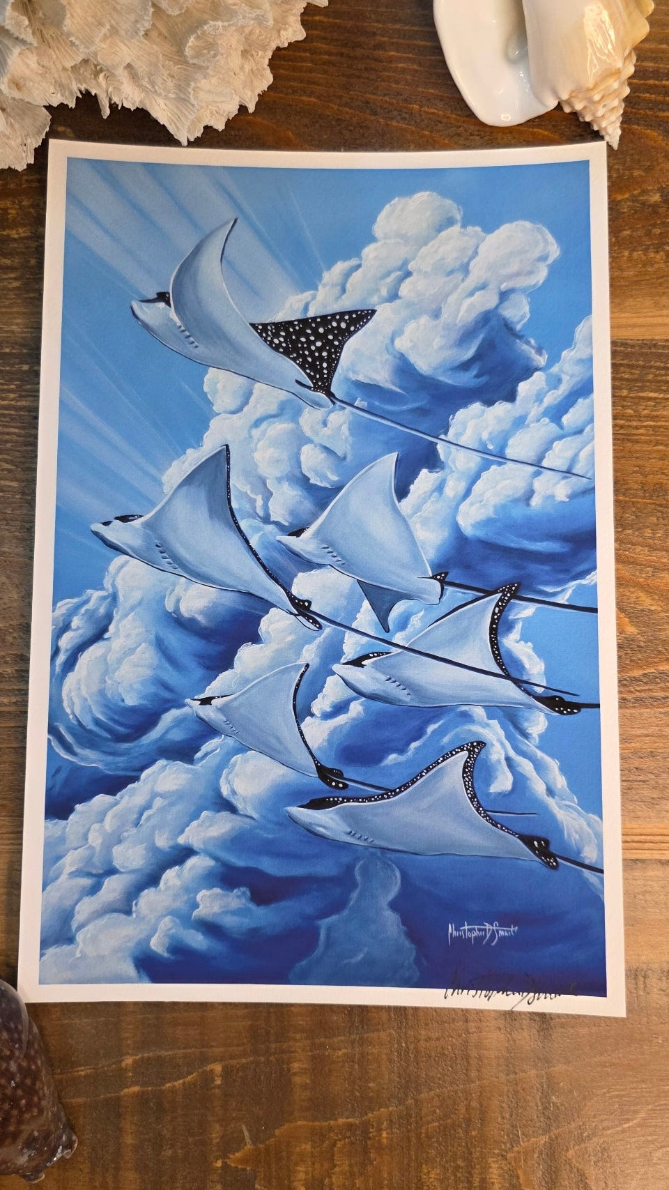 A square art print depicting a school of manta rays swimming through blue ocean waves, styled on a wooden surface with seashells nearby.