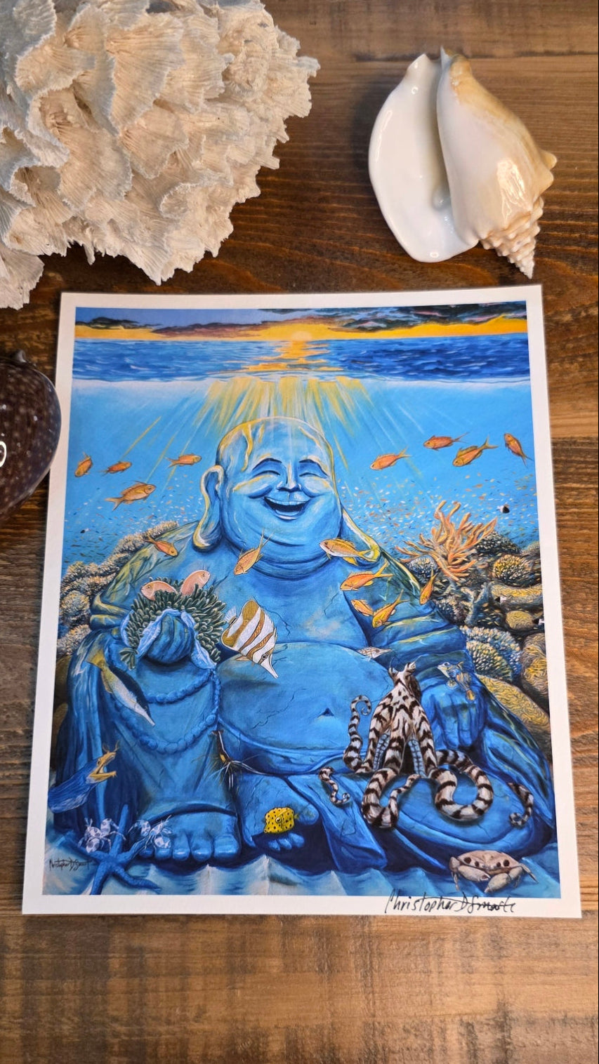 A square art print featuring a blue Buddha illustration with ocean-themed details, displayed on a wooden surface with seashells and coral around it.
